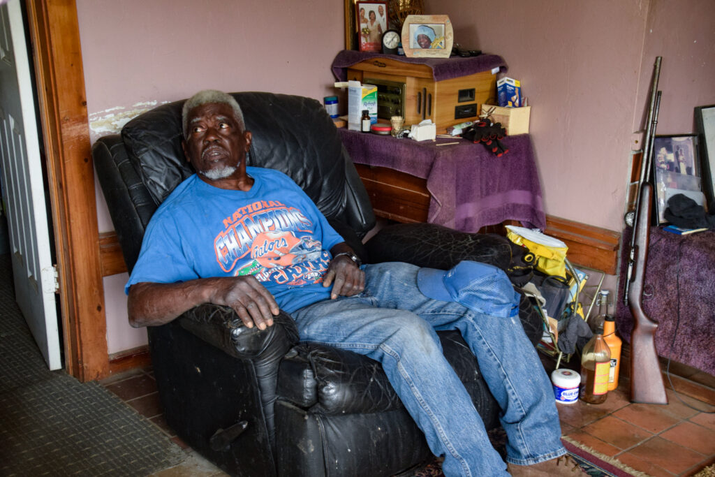 A Black man in a blue shirt and blue jeans rests his blue hat on his knee as he sits back in his black leather recliner. Surrounding him is a gun, picture frames and a variety of random house items like bandaids, gloves and a mini clock.