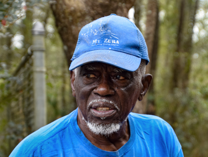 A Black man in a blue "Mt. Zura" hat and blue shirt looks off into the distance.