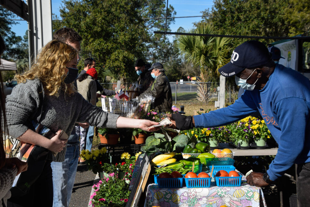 A blonde-haired girl in a black mask hands a Black man in a blue surgical mask a five dollar bill. They are surrounded by red tomatoes, green peppers, yellow squash, pink flowers and other people wearing masks at a farmer's market.