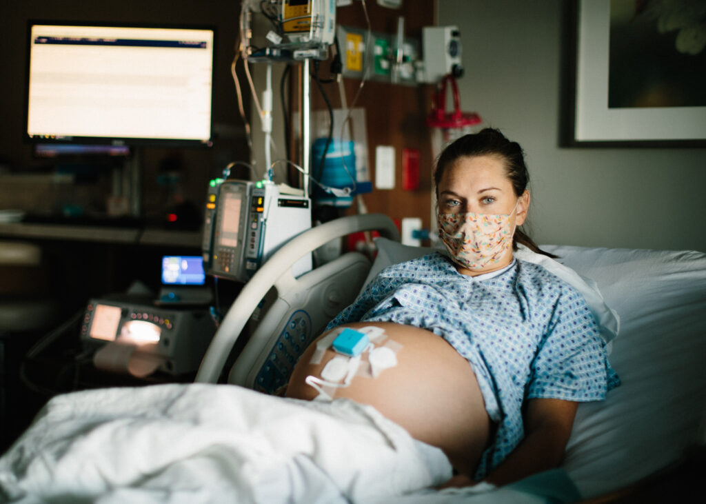 A pregnant woman wearing a mask lays back on a hospital bed with various monitors connected to her belly