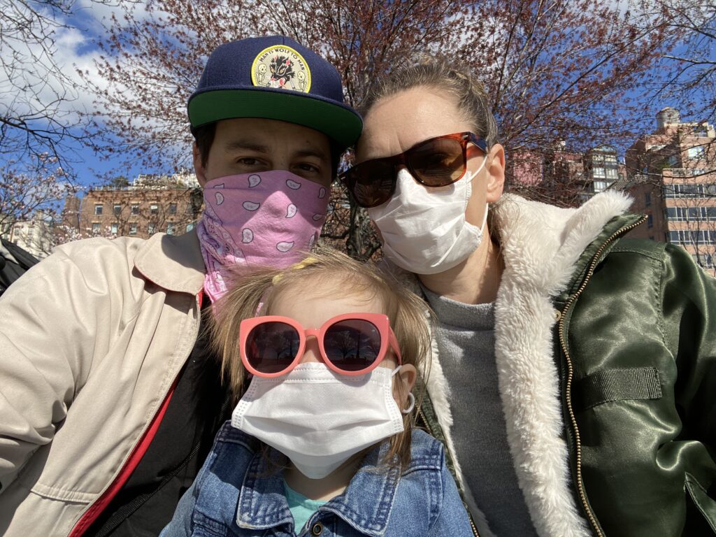 A family including a man, woman and their toddler daughter take a selfie while wearing masks and sunglasses