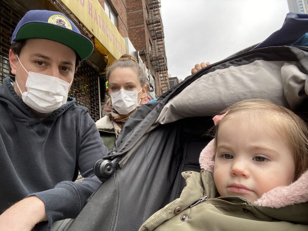 A man and woman wearing masks crouch next to their toddler daughter in her stroller with city buildings in the background