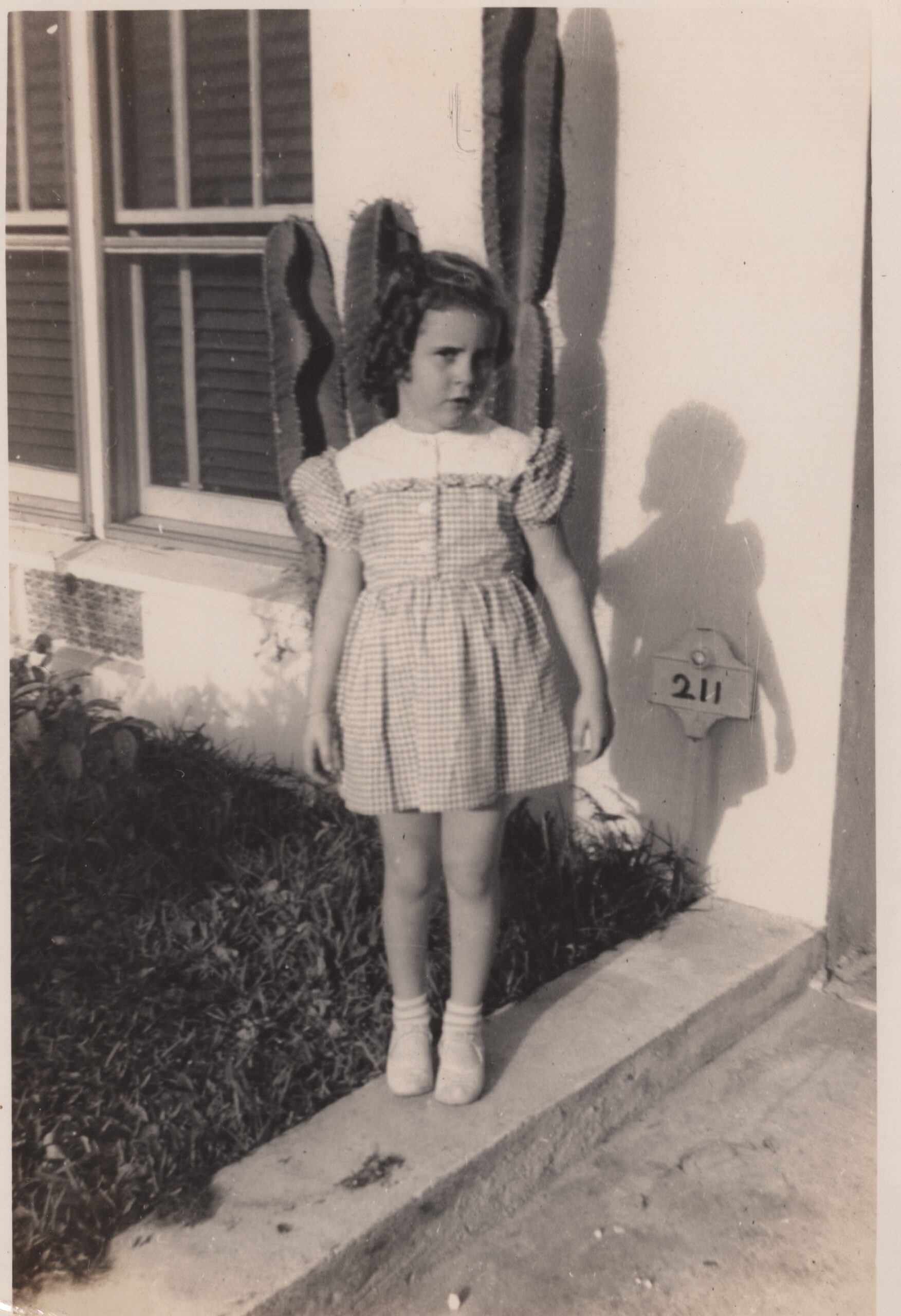 Judy, about five or six, stands with her arms at her side. She wears a gingham dress and Mary Jane-style shoes. Her hair is worn is tight ringlets above her shoulders.